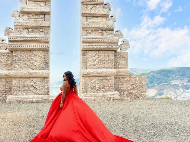 VickyGreys with big ass, wearing a red ball gown, standing in front of ancient stone pillars, blue sky, and mountains in the background. Dark skin, black wavy hair.