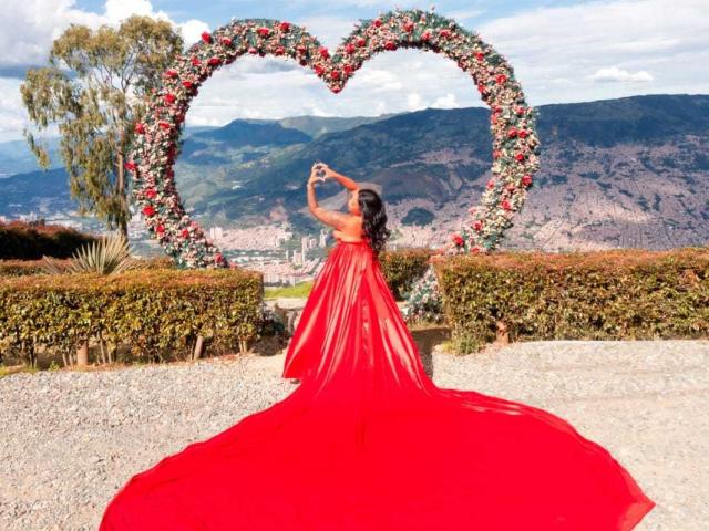 VickyGreys with red, floor-length, sheer gown, standing in front of heart-shaped flower arrangement, arms raised, long, wavy hair, facing away, mountain landscape in background.