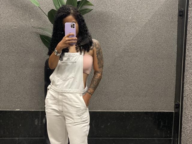 Meel69 with curly black hair, wearing white overalls, pink bra showing tits, tattoos on right arm, holding purple phone, standing in front of gray wall, black floor.