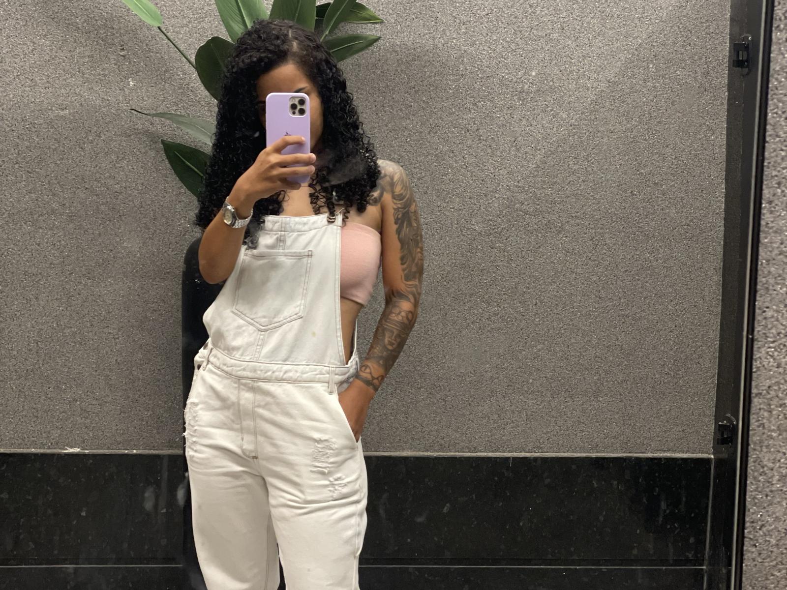 Meel69 with curly black hair, wearing white overalls, pink bra showing tits, tattoos on right arm, holding purple phone, standing in front of gray wall, black floor.
