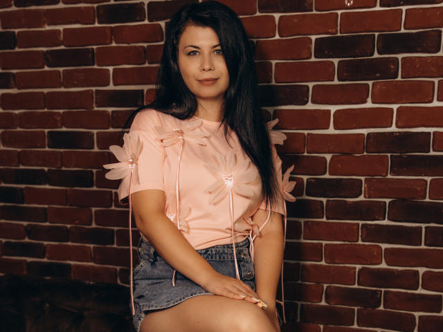CharloteNoir with long black hair, pink floral shirt, and denim skirt, sitting against a brick wall, showing a cute smile, with small tits and a sexy, relaxed look.
