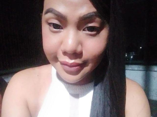 TsHOTGODDESS-hot with long black hair, brown skin, and a cute smile, wearing a white sleeveless top, looking down with subtle eyeliner. Boobs not visible, just her face.