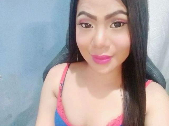 TsHOTGODDESS: una mujer atractiva con cabello negro largo, delineador de ojos rosa y lápiz labial rosa, vestida con una blusa de encaje rosa y azul, mirando a la cámara con una ligera sonrisa..