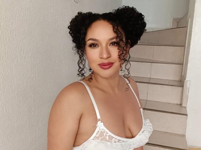 CarlaMuille with curly black hair, wearing a white lace bra, showing her tits, smiling, standing in front of a staircase. She's got red lipstick and a cute face.