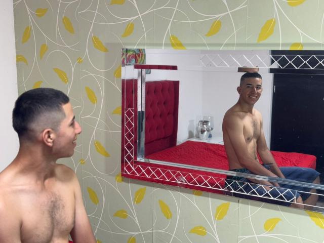 MartinBlos is shirtless with a shaved head, sitting in front of a mirror, reflecting his bare chest and a tattoo on his right arm, wearing blue jeans.