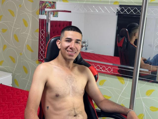 MartinBlos, shirtless, sitting in a black and red chair, smiling, short dark hair, light skin, small tattoo on right side, mirror reflection showing his back, red bed in background.