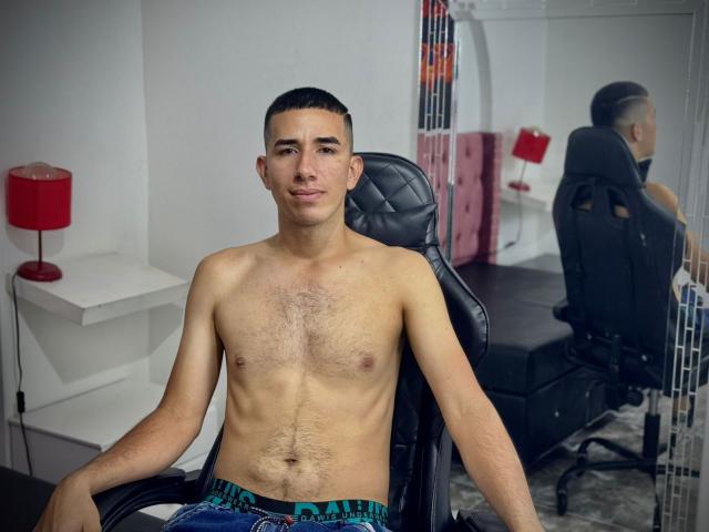 Shirtless MartinBlos with a slim, hairy chest, sitting in a black chair, wearing blue and green patterned shorts, short dark hair, and a slight smile, in a room.