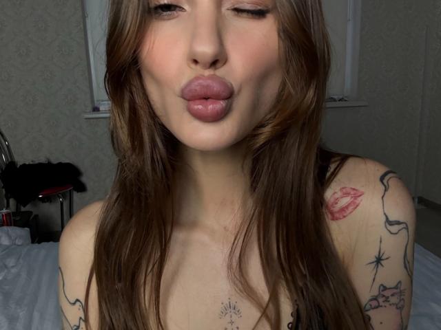 SensualMila with pouty pink lips, brown hair, tattoos on shoulders and chest, topless, bedroom, soft light. Boobs visible, tattoos include red lips, black lines, star.