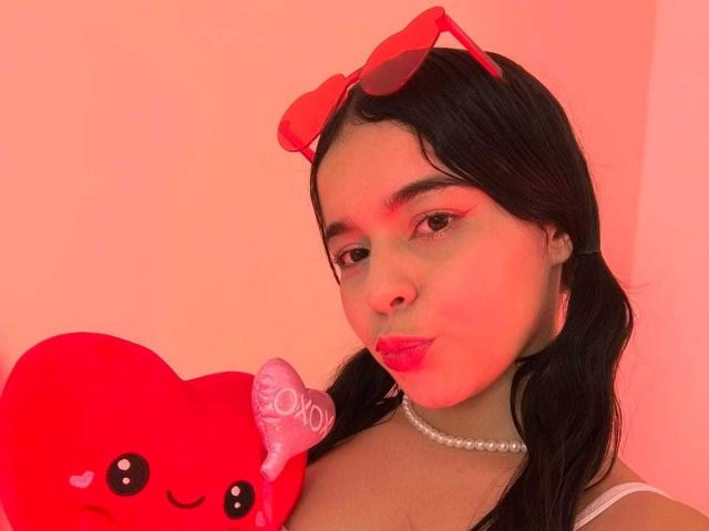LissyCarter with black hair, red eyeshadow, pink lipstick, pearl necklace, red heart stuffed animal, and red sunglasses on her head, puckering her lips. Pink lighting.