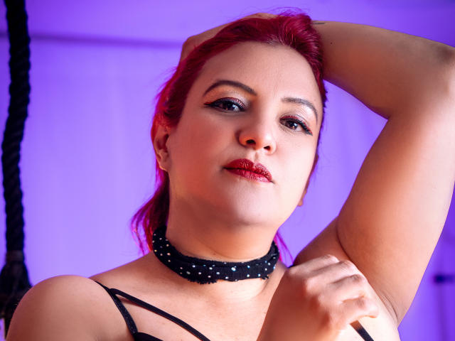 SammyOwens met rood haar, zwarte eyeliner, rode lipstick, een zwarte choker, en een arm omhoog, die haar borsten laat zien in een zwarte top, tegen een paars verlichte achtergrond..