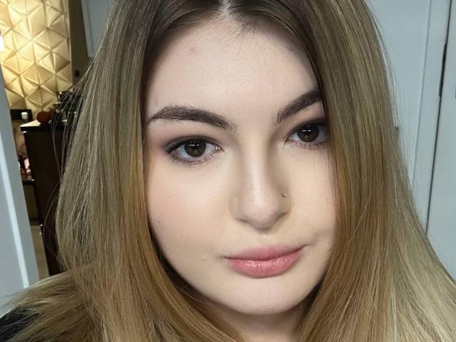 CattieHolly has light brown eyes, straight blonde hair, a small nose piercing, and pink lips. Her eyebrows are dark and she's looking slightly to the right.