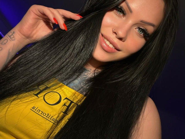 KittyMissyX with long black hair, red nails, nose ring, tattoo on neck, smiling, green eyes, wearing yellow top, holding hair, visible blue light in background.