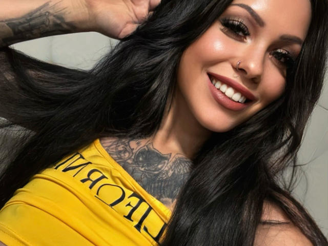 KittyMissyX with long black hair, smiling, yellow top, nose ring, tattooed chest, and visible tits, holding hair, shiny makeup, and full lips.