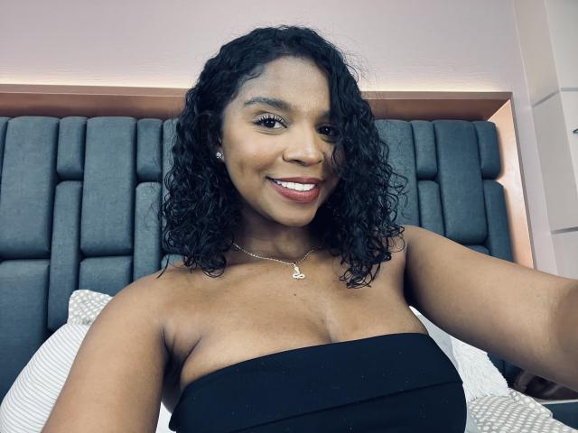 LisaMendez with curly black hair, dark skin, smiling, wearing a strapless black top, showing her boobs, necklace with heart pendant, in a bedroom, taking a selfie.