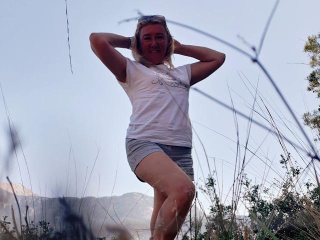 KarolinaInsky with blonde hair, white t-shirt, and gray shorts, standing with hands behind head, smiling, in grassy field, legs crossed, sunlight on face, mountains in background.