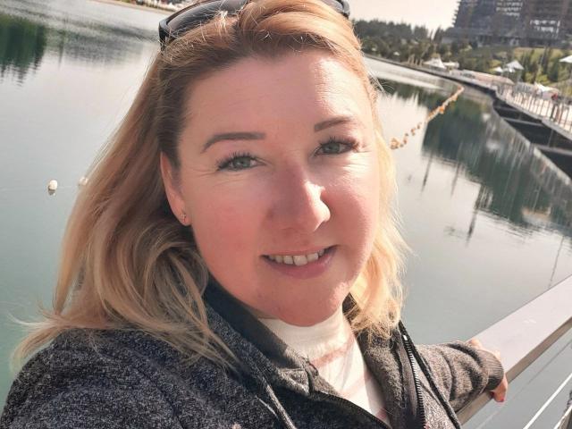 KarolinaInsky has blonde hair, green eyes, and a smile, wearing a gray jacket with sunglasses on her head, standing by a reflective waterway, her hand on a white railing.
