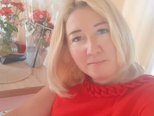 KarolinaInsky with blonde hair, blue eyes, and red dress, looking at camera with slight smile, red flowers in glass vase to left, black object behind.