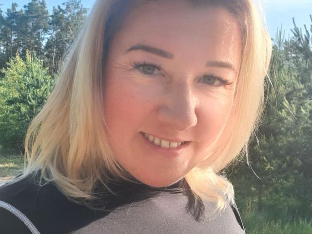KarolinaInsky with blonde hair, fair skin, and a cute smile, wearing a black top, has noticeable tits, and is standing outdoors in sunlight.