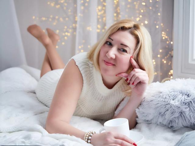 KarolinaInsky with blonde hair, white knit dress, and pearl bracelet, lying on white bed, holding white mug, smiling, legs up, fingers touching cheek, red nails, soft lighting.