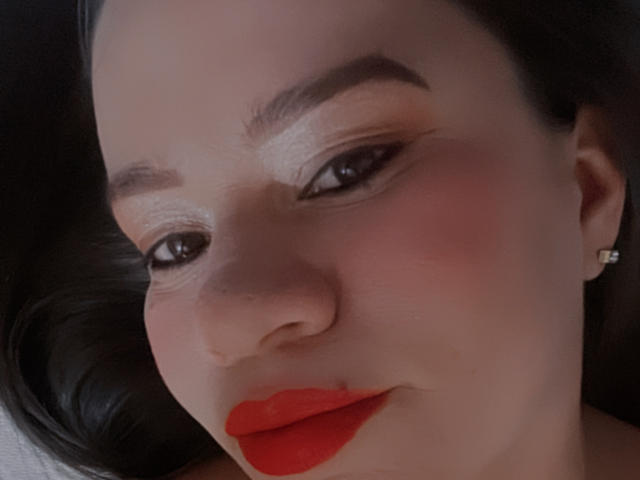 Het gezicht van CharllotI, met rode lippenstift, zwarte eyeliner en rode blush op haar lichte huid, met donkere wenkbrauwen en bruine ogen, liggend met haar zwarte haar gedeeltelijk zichtbaar..