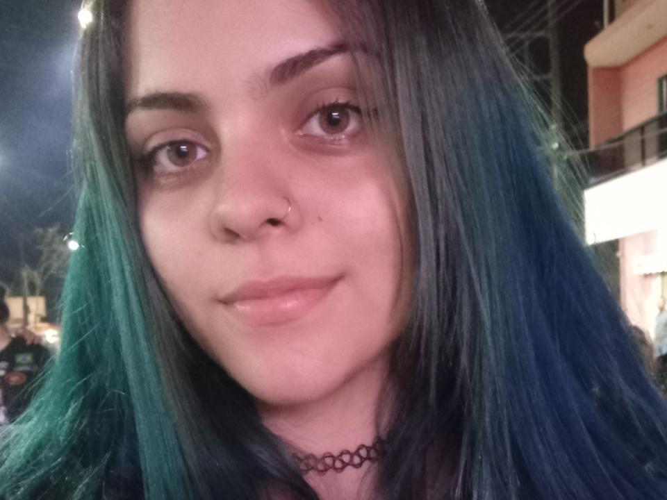 HotDocinho has green-blue hair, a nose ring, and a slight smile with pinkish lips. She's wearing a black choker and looking at the camera with brown eyes.
