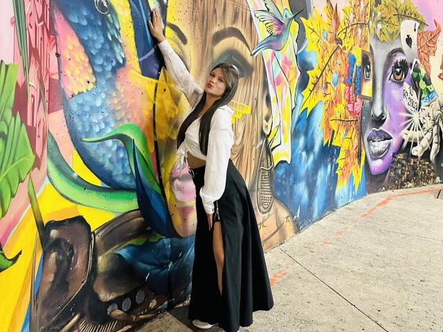 VictoriaWaynes with long black hair, white blouse, black high-waisted skirt, holding pink ice cream, leaning against colorful graffiti wall, showing her ass, standing on concrete.