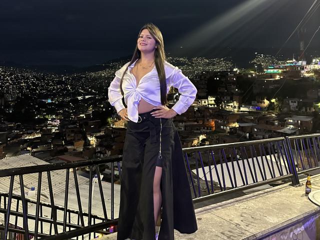 VictoriaWaynes with long brown hair, white tied-up shirt showing her tits, black high-waisted skirt with a leg slit, standing on a rooftop at night, smiling.