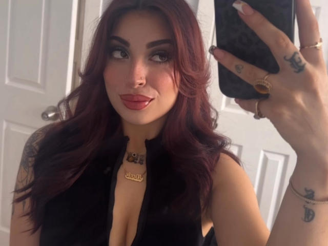 MaddyCalloway with red hair, black sleeveless top, cleavage, and tattoos, taking a selfie with a smartphone, showing off a ring and necklace.
