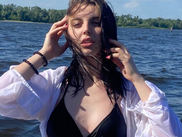 AliceJayms with wet black hair, wearing a white shirt and black bikini top, hands in her hair, blue water, and green trees in the background, sunlight on her face.