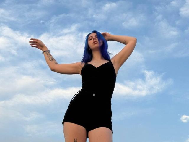 AliceJayms with blue hair, black shorts, and black top, arms raised, tattoo on left arm, standing against a cloudy sky. Her ass is prominent, with a small tattoo on her right thigh.