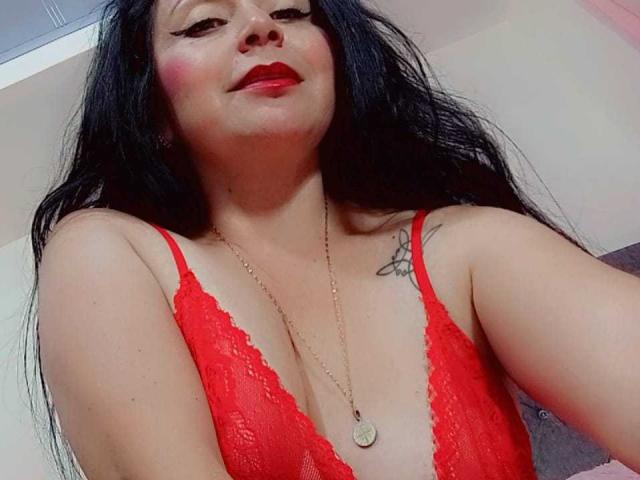 Zaria69, com cabelos pretos longos, sutiã de renda vermelho, batom vermelho, colar de ouro, uma pequena tatuagem no ombro direito, sorrindo, mostrando os seios. Sua pele é morena clara..