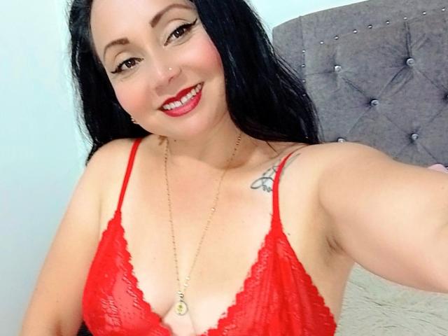 Zaria69 with black hair, red lace bra, smiling, showing tits, gold necklace, tattoo on right shoulder, holding phone for selfie.