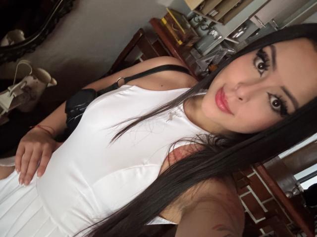 AlisonTatcher with long black hair, wearing a white dress, black strap, and pink lipstick, holding a black bra strap, smiling at the camera, showing her tits.