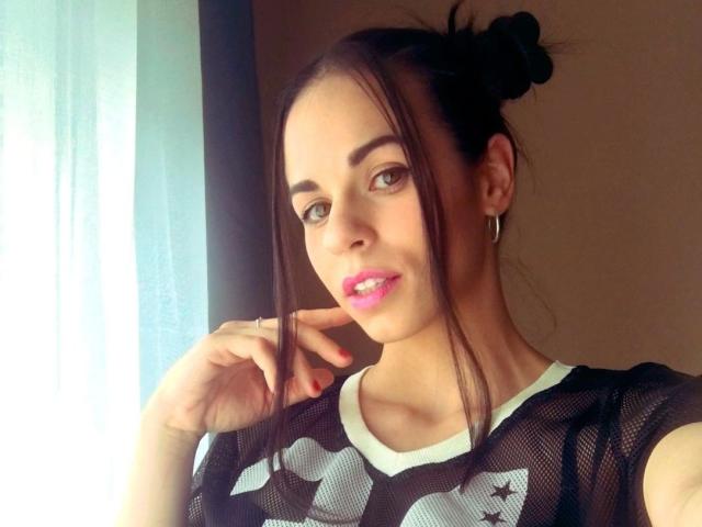 Nekane-hot has pink lipstick, brown eyes, and her dark hair in a messy bun. She's wearing a black mesh shirt with white and gray patterns, showing her tits.