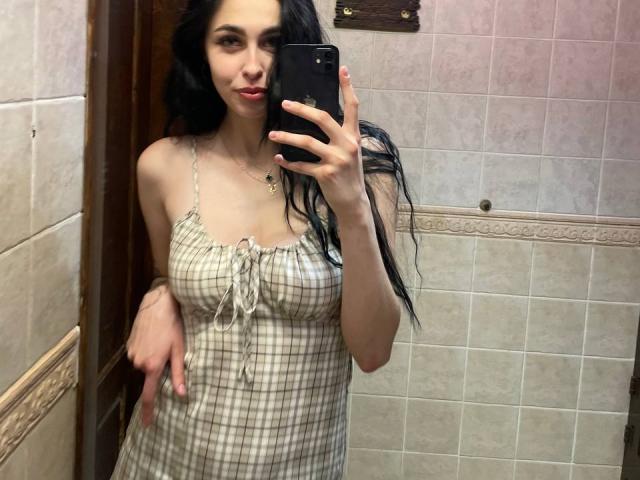 Luyizia with long black hair, wearing a checkered dress, holding a phone, showing her tits, standing in a tiled bathroom, smiling slightly, with one arm raised.