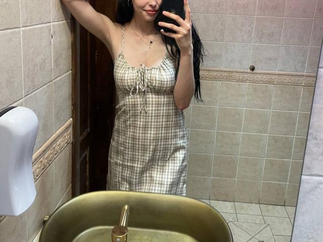 Luyizia with long black hair, wearing a checkered dress, holding a phone, standing in a bathroom, showing her tits, in a mirror selfie, holding a phone.