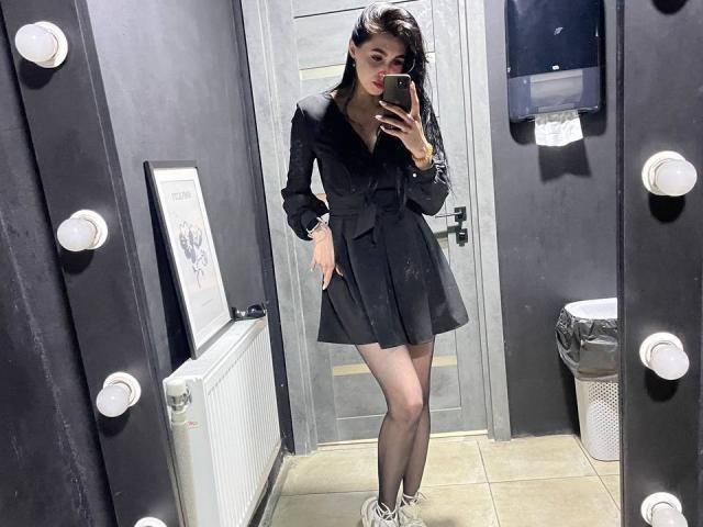 Luyizia indossa un abito con colletto in pelliccia nera, una gonna nera, collant trasparenti e scarpe da ginnastica bianche, e si sta scattando un selfie allo specchio in un camerino illuminato, mostrando il sedere e i piccoli seni..