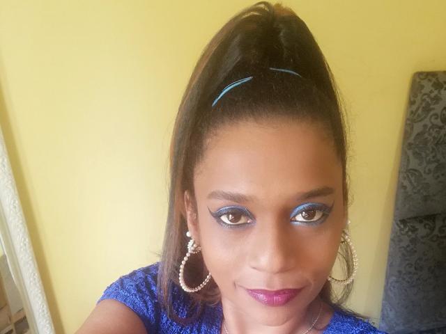 MiZzCreator with dark brown eyes, blue eyeliner, and purple lipstick, wearing a blue lace top, hoop earrings, and a high ponytail, smiling at the camera.