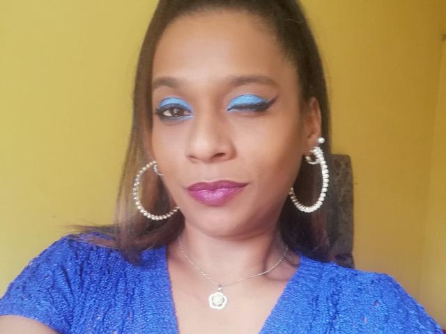 MiZzCreator has blue eyeliner, pink lipstick, hoop earrings with pearls, a necklace, and a blue textured top. She's smiling with brown hair in a ponytail.