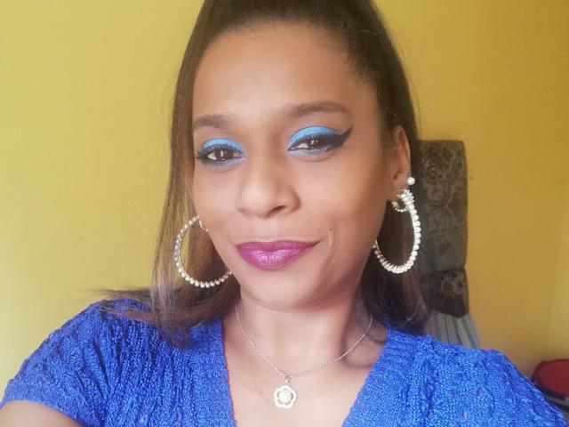 MiZzCreator has blue eyeshadow, shiny pink lipstick, hoop earrings with pearls, a blue textured shirt, and a necklace with a circular pendant, looking cute and confident.