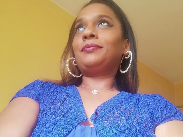 MiZzCreator has medium brown skin, blue eyes, and wears a blue knit top with a keyhole cutout, hoop earrings, and a necklace. She's looking up with a slight smile.