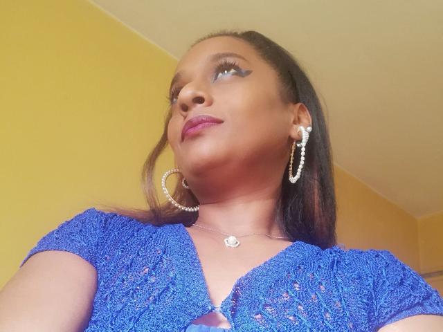 MiZzCreator with dark skin, purple lipstick, blue crochet top, hoop earrings, and pearl necklace, looking up with eyeliner accentuating her eyes. Boobs visible through top's cutout.