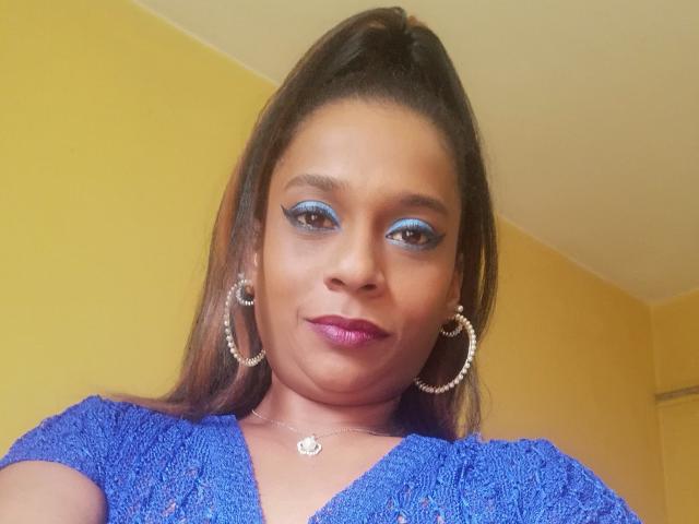 MiZzCreator with blue eyeshadow, purple lipstick, hoop earrings, and a blue textured dress, showing a confident smile, brown ponytail, and a silver necklace.