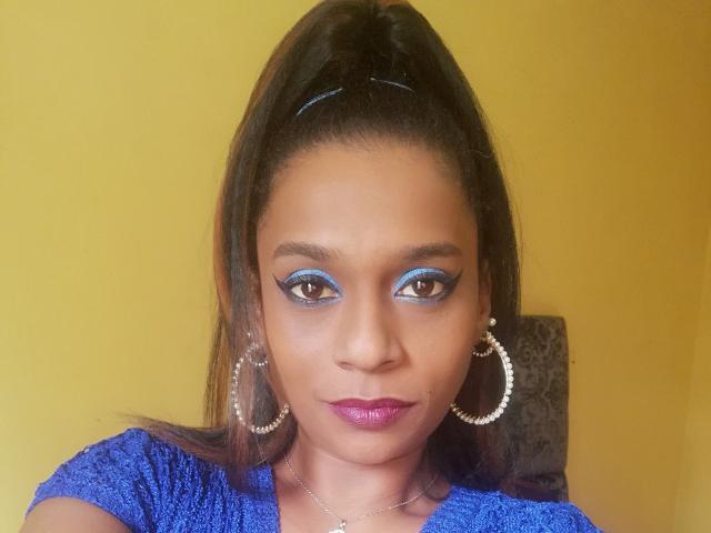 MiZzCreator has dark brown eyes with blue eyeliner, shiny purple lipstick, and wears large silver hoop earrings, a blue textured shirt, and a high ponytail.