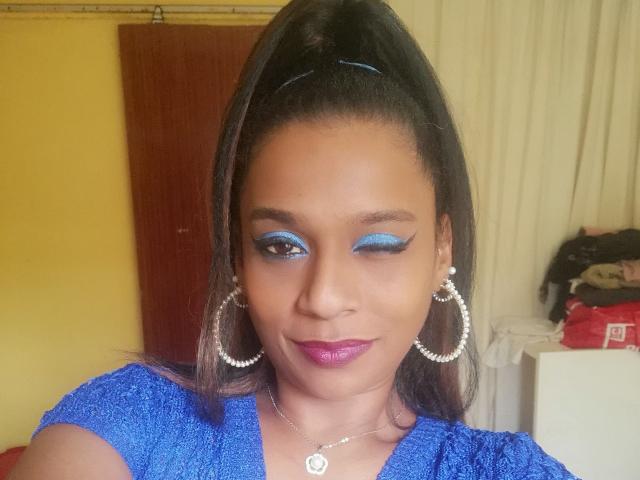 MiZzCreator with dark brown skin, blue eyeshadow, purple lipstick, blue top, high ponytail, silver hoop earrings, and necklace, smiling at the camera. Boobs not visible.