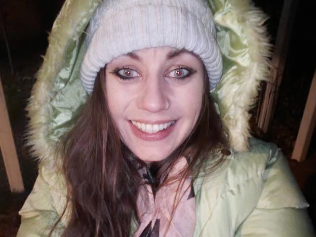 MissmeanDre with brown eyes, white knit beanie, green hooded jacket with fur trim, smiling, showing white teeth, long brown hair, wearing pink shirt, nighttime selfie.