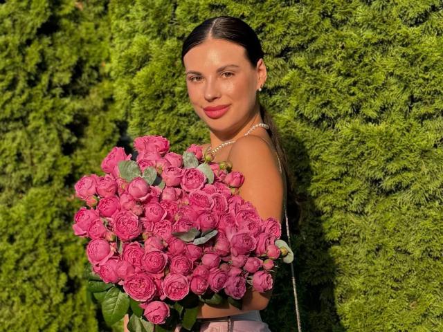 BlackFatal met donker haar, roze rozen, die een grote bos bloemen vasthoudt, draagt een roze top, een parelketting, glimlacht, staat voor een groene haag, zonnig, schattig..