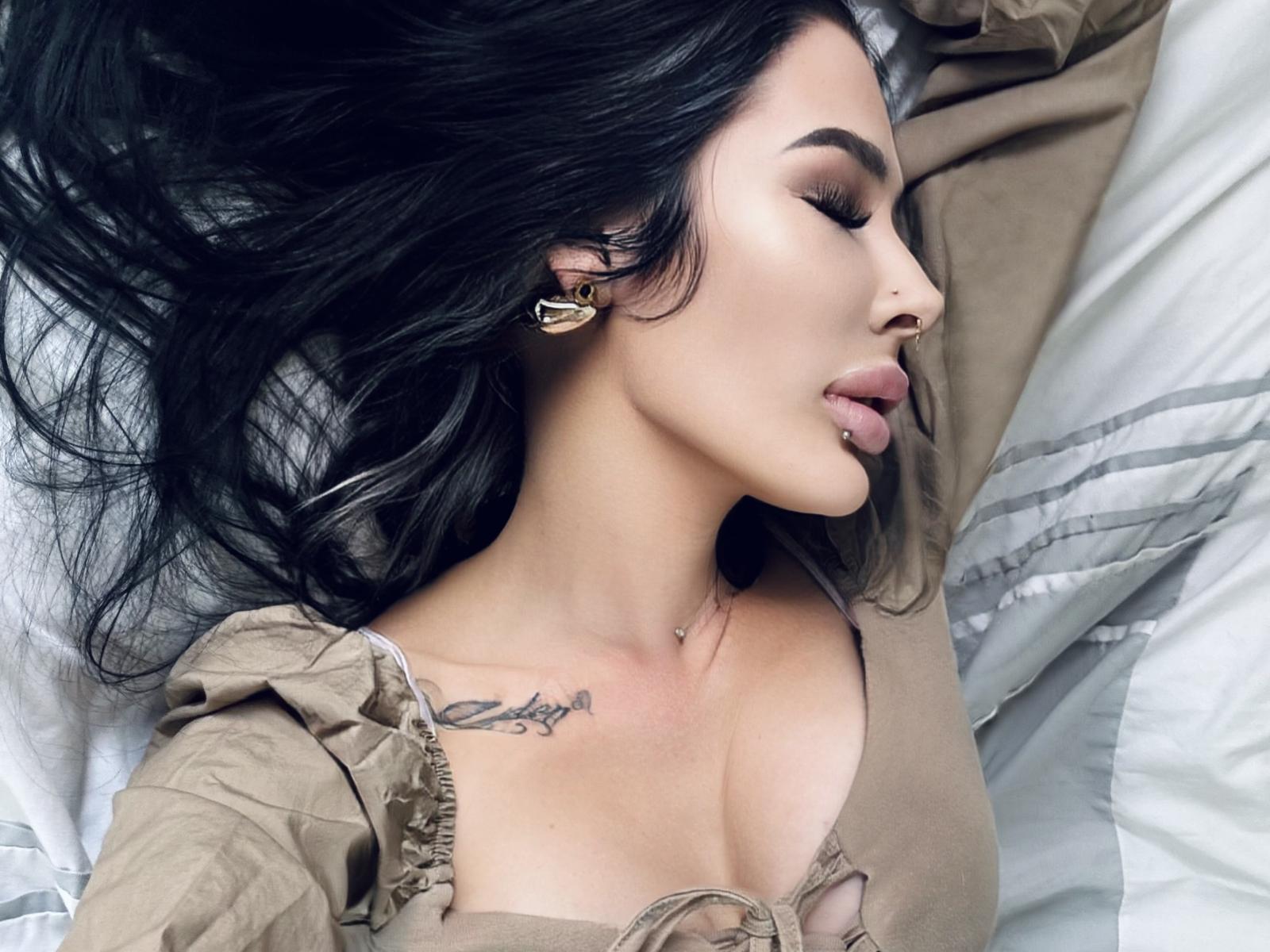 TheLittleGina's face is in profile, black hair spread, gold earrings, tongue out, tan top, visible tits, tattoo above, nose ring, and light makeup, lying on a white pillow.