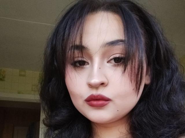 MyrkaHot has dark, wavy hair, full red lips, and a small beauty mark below her right eye. Her makeup is subtle, with dark, defined eyebrows.