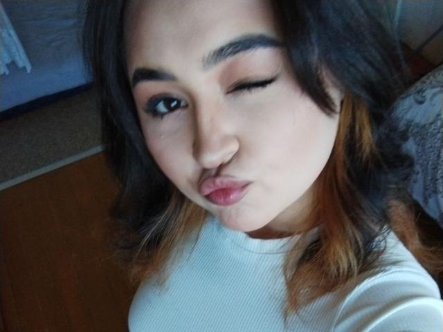 MyrkaHot has wavy dark brown hair with orange highlights, one eye closed, and puffed lips, wearing a white ribbed shirt. Her eyebrows are thick and she's winking.